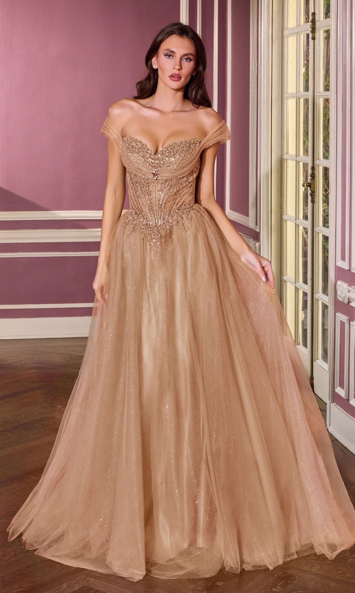 A woman wearing the Ladivine CC493 long prom dress in mocha gold, featuring an off shoulder sweetheart neckline and intricate beading, stands in a room with mauve walls and white trim.